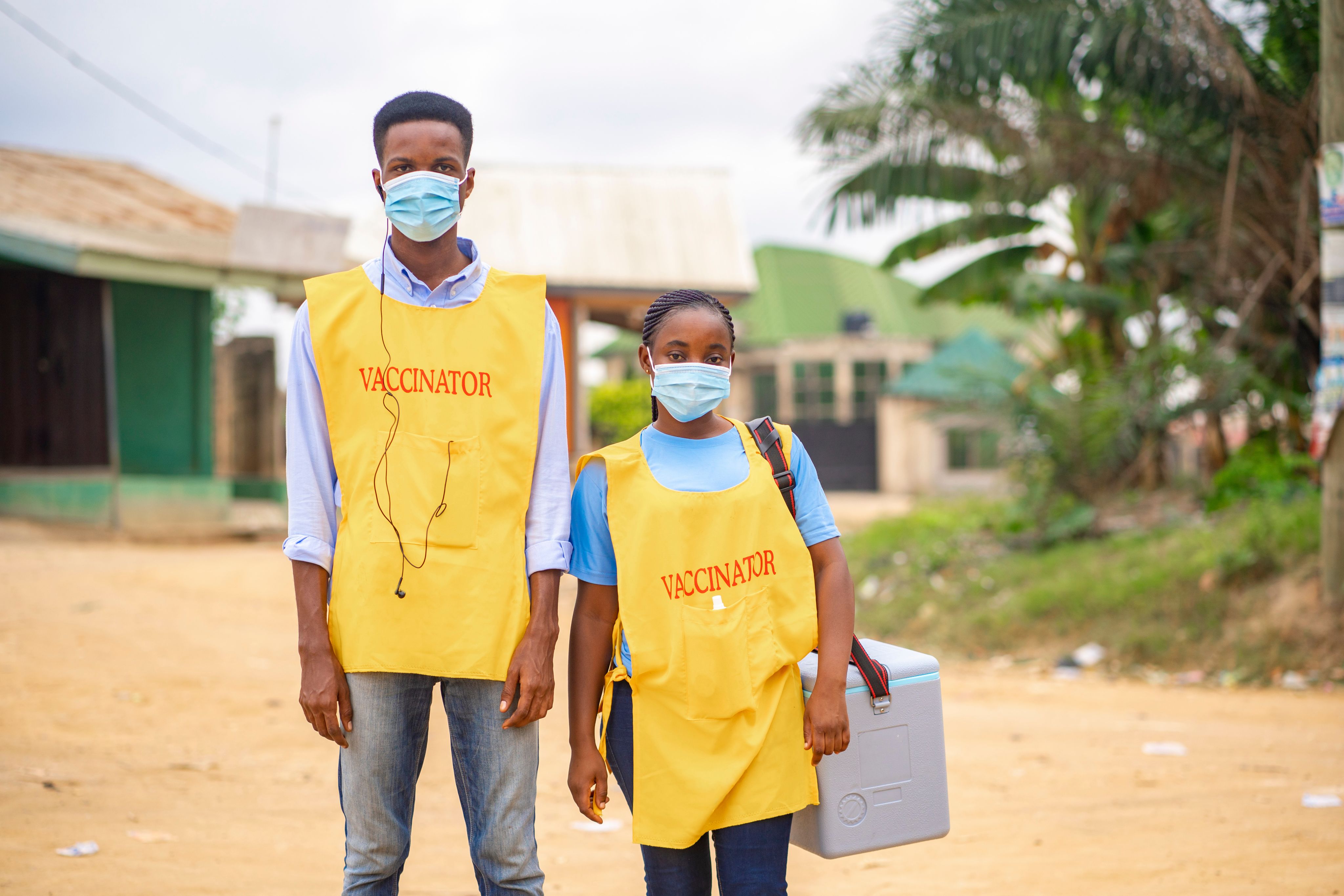 Two health workers with bibs with VACCINATOR written on them stand in a remote village